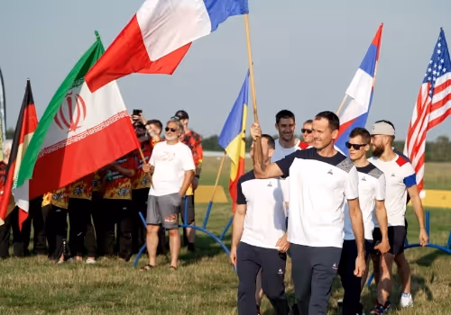 Skupina mužů kráčí na louce, někteří nesou vlajky Francie, Německa, Íránu, Ukrajiny, Srbska a USA.